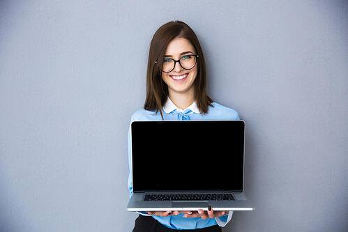 Happy businesswoman showing blank laptop screen over gray background. Wearing in blue shirt and glasses. Looking at camera
