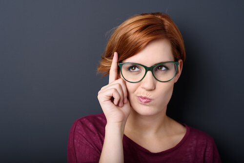 Nerdy scholastic young woman wearing geeky glasses standing thinking with her finger raised and a grimace of concentration in a humorous stereotypical depiction, over a dark background with copyspace-1