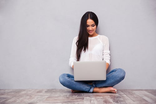 Portrait of a happy casual woman sitting on the floor with laptop on gray background